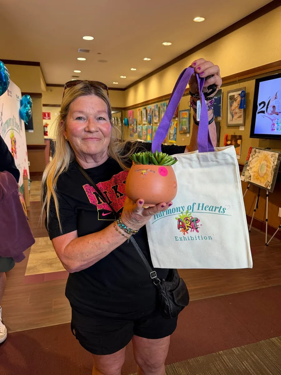 A smiling visitor holds up a potted plant and a Harmony of Hearts tote bag.