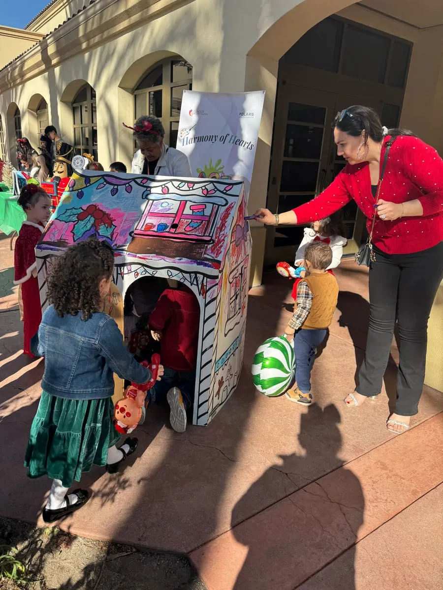 Children color a giant cardboard house outdoors as a volunteer helps.