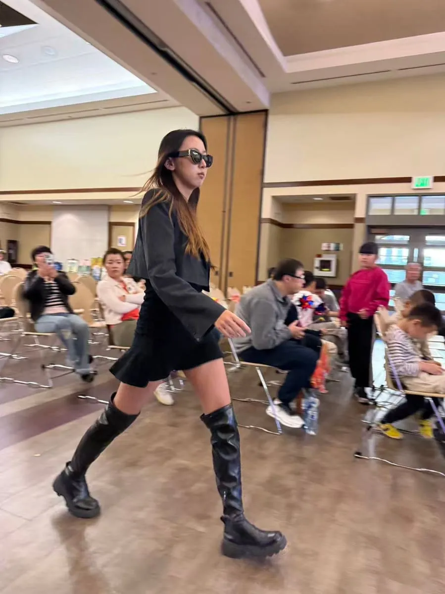 A girl in sunglasses and boots walks the runway at a kids’ fashion show.