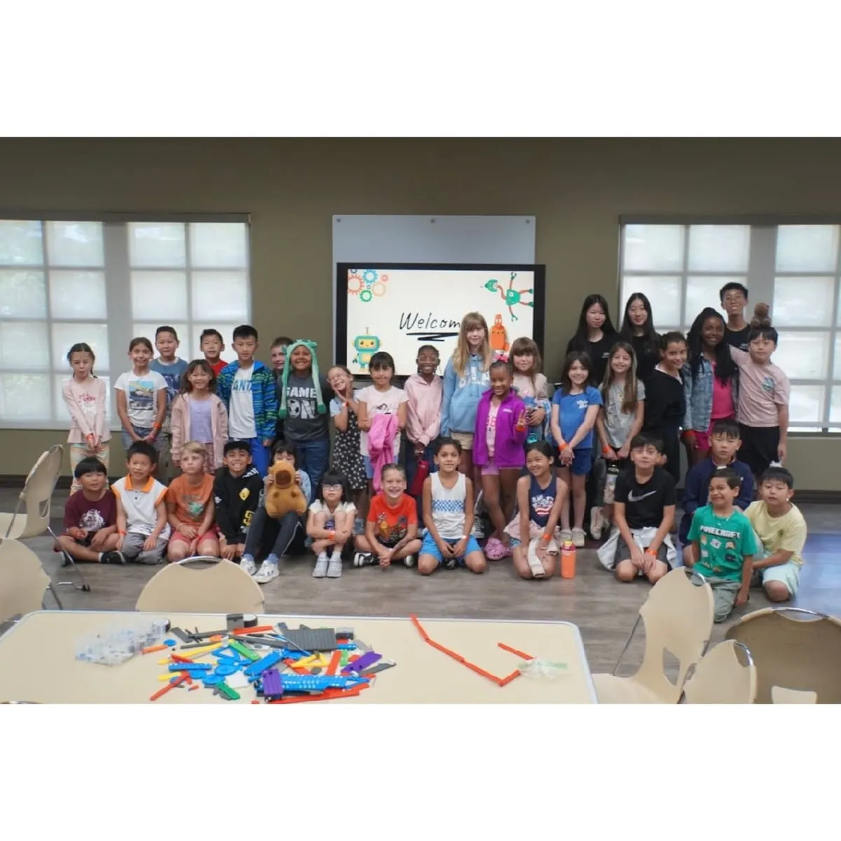 A large group of children gathers in front of a “Welcome” screen with robot graphics.