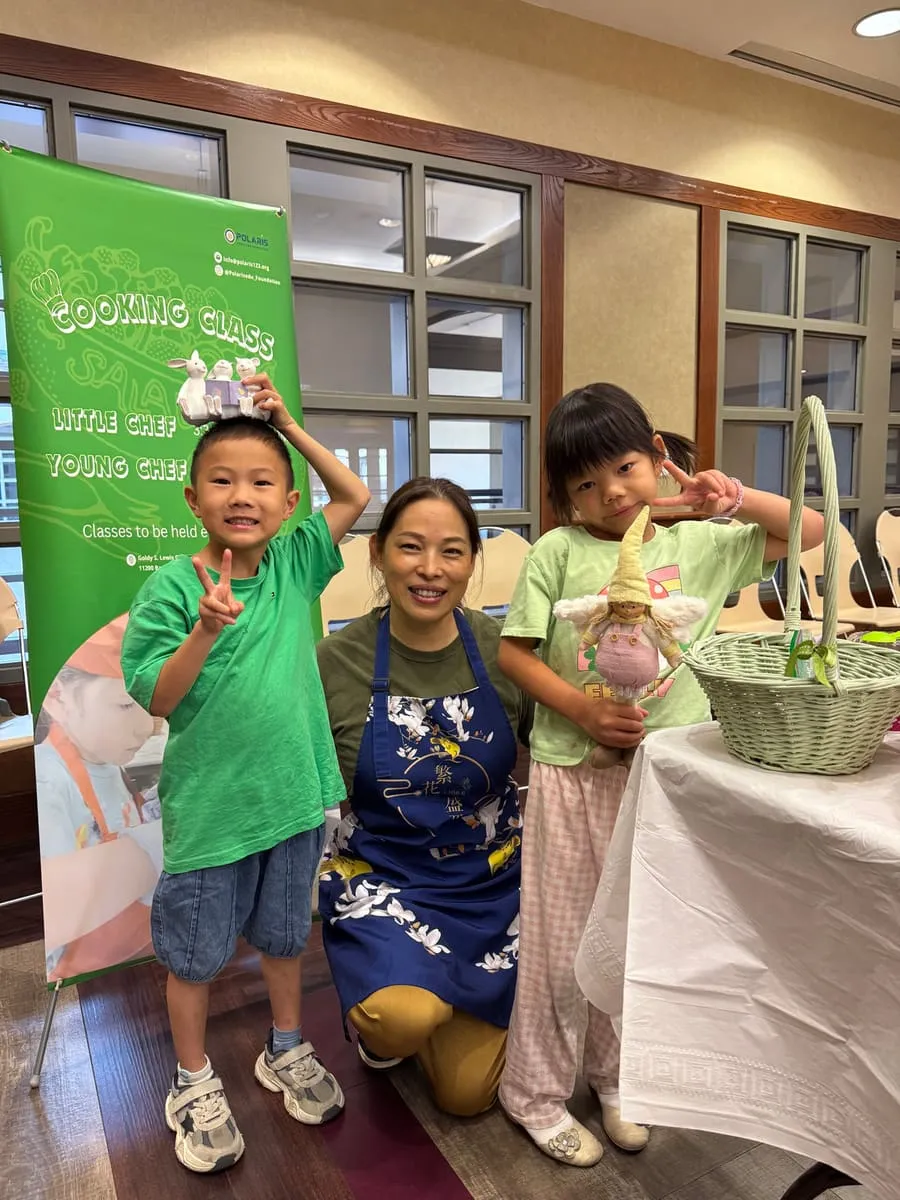 An instructor kneels with two children beside the Cooking Class banner.