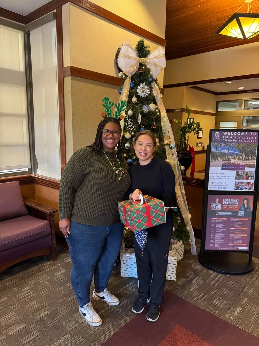 Two women hold a wrapped gift beside a Christmas tree.