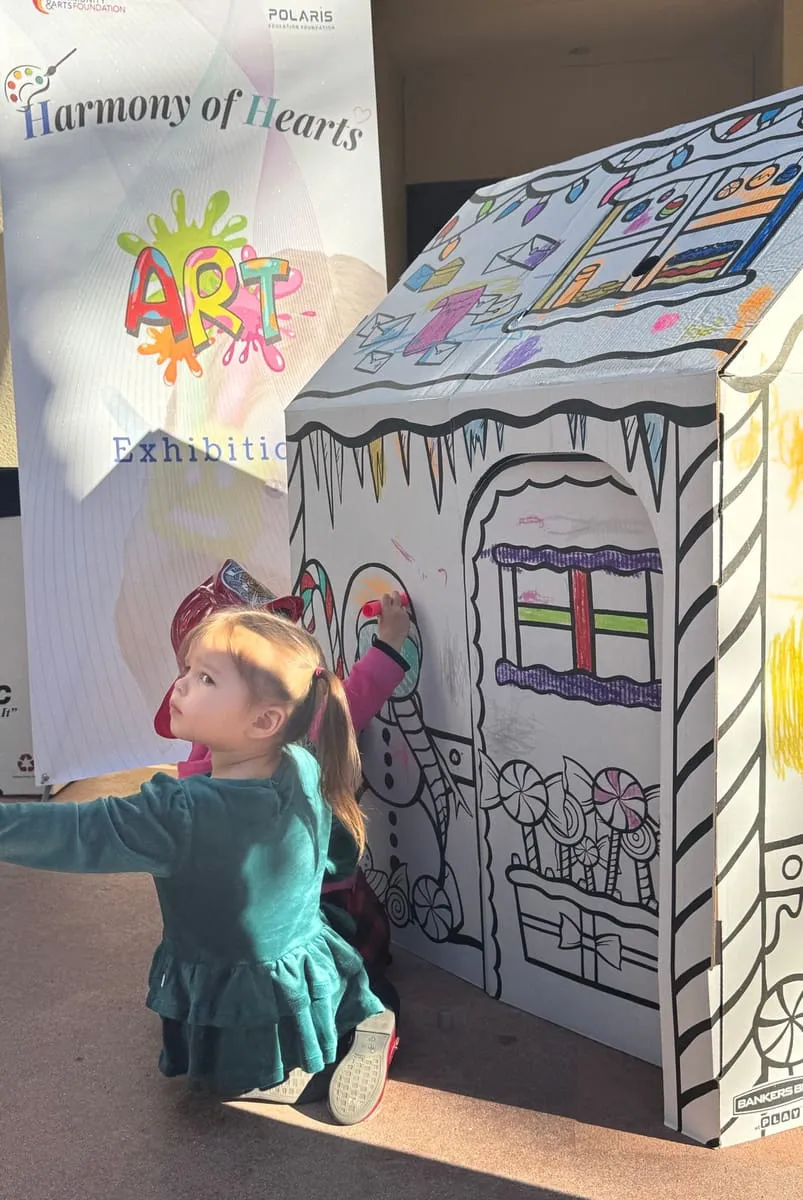 A toddler colors a cardboard house beside a Harmony of Hearts banner.