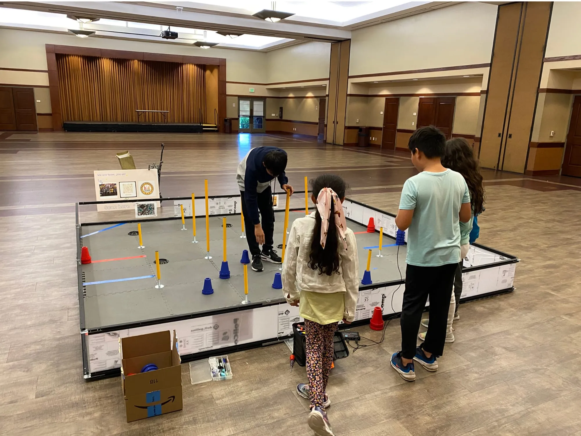 Students set out cones and poles on a robotics competition field in a community hall.
